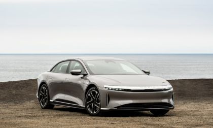 A silver Lucid Air luxury electric sedan shown from a front three-quarter angle, parked on a coastal terrain with the ocean visible in the background.