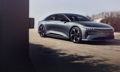 A gray 2025 Lucid Air Touring is parked on sandy terrain, framed by beach grass and wooden posts, showcasing its modern design.