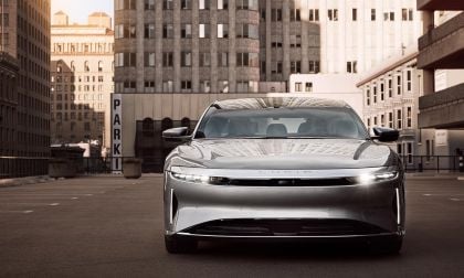 A silver 2025 Lucid Air parked in an urban setting, surrounded by tall buildings and a "PARK" sign in the background.