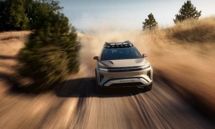 Silver Lucid Gravity electric SUV shown in dynamic three-quarter front view, kicking up dust while driving on dirt road, featuring distinctive LED lighting and roof rack.