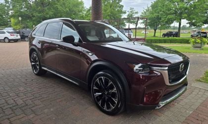 Deep burgundy 2025 Mazda CX90 SUV parked on brick pavement, angled front three-quarter view with lush green trees in background.