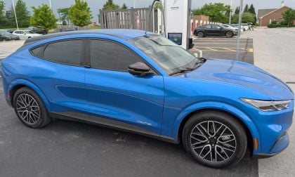 A blue Ford Mustang Mach-E parked near an electric vehicle charging station, surrounded by trees and other cars.