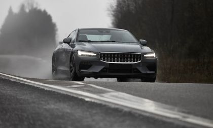 A sleek gray car drives through a misty landscape, splashing water on a wet road, showcasing its dynamic design and performance.