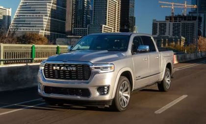 Silver 2025 RAM 1500 pickup truck driving on a city bridge with modern skyscrapers in the background. The scene conveys a sense of urban adventure.