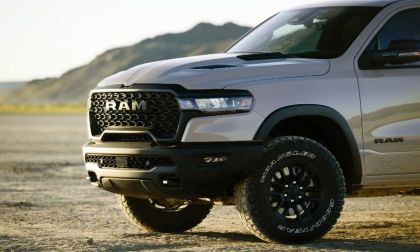 A silver 2025 RAM 1500 pickup truck shown from the front quarter angle, featuring black off-road wheels, aggressive honeycomb grille design, and rugged front bumper against a mountainous backdrop.