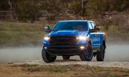 A bright blue 2025 Ram 1500 pickup truck shown from a front three-quarter angle, with LED headlights illuminated and off-road tires kicking up dust on rugged terrain.