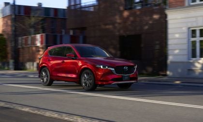 A 2025 red Mazda CX-5 cruising down a city street, showcasing modern architecture and vibrant colors in the background.