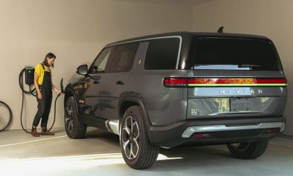 A gray Rivian SUV is parked in a garage, connected to an electric charger, while a person stands nearby, holding the charging cable.