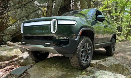 A dark green Rivian R1S electric SUV shown from the front quarter angle, demonstrating its off-road capabilities while navigating rocky terrain in a wooded setting.