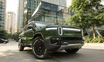 Low-angle view of a green 2025 Rivian R1S electric SUV driving through a modern city street with glass buildings
