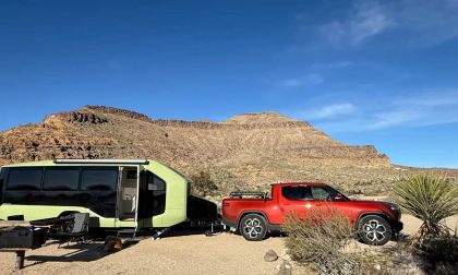 A red Rivian R1T pickup truck is parked next to a lime green camper trailer in a desert landscape with rocky mountains in the background, photographed from a side angle.