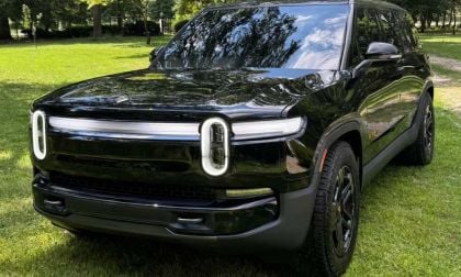 A sleek black electric SUV with distinctive round headlights and a glossy finish, parked on a grassy area surrounded by trees.