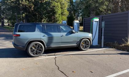A silver 2025 Rivian R1S is shown from the rear three-quarter view at an EV charging station, featuring black accent wheels and trim.