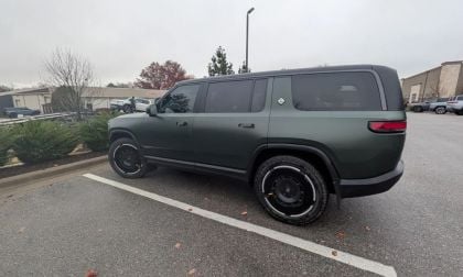 Forest Green 2025 Rivian R1S SUV photographed from a rear three-quarter angle in a parking lot, showing its boxy design, black wheels, and matte finish.