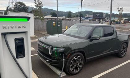 Dark green Rivian R1T electric pickup truck charging at a white Rivian charging station in an overcast suburban parking lot with mountains in background