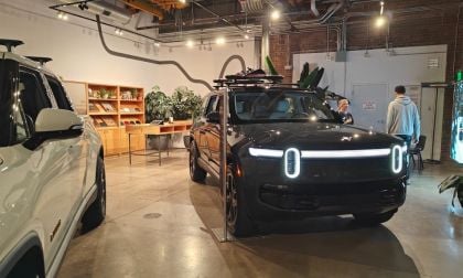 2025 Rivian R1S electric SUV in black, front three-quarter view, displaying distinctive LED headlight bar, shown in industrial-style showroom with exposed brick walls.