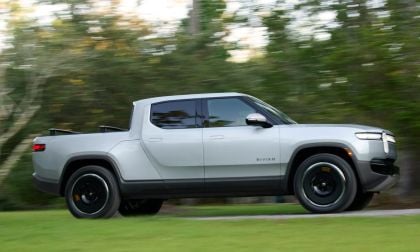Photograph of the 2025 Rivian R1T electric truck in a gray color, captured from a three-quarter front angle with trees visible in the background.