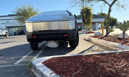 Tesla Cybertruck parked in a commercial parking lot with a large white boulder lodged under its front end, showing the electric truck elevated slightly off the ground.