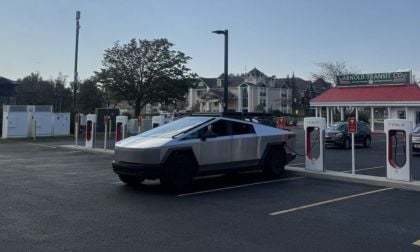 A Tesla Cybertruck charging at a station, surrounded by other chargers and vehicles, set against a backdrop of trees and buildings.