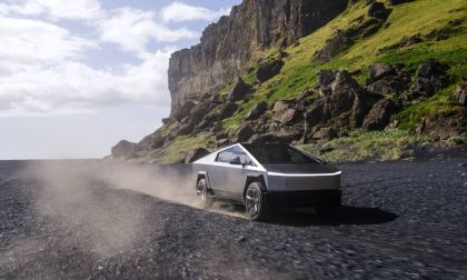 A silver Tesla Cybertruck shown from a rear three-quarter angle, kicking up dust while driving on a dramatic coastal road with steep cliffs in the background.