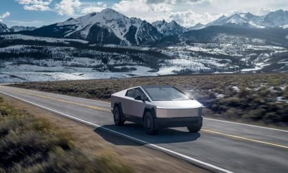 A futuristic electric truck drives along a scenic road with snow-capped mountains and a clear blue sky in the background.