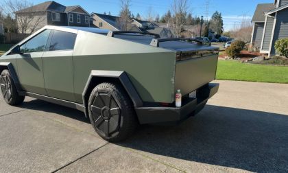 A matte gray Tesla Cybertruck shown from a rear three-quarter angle, featuring its distinctive angular design and large geometric wheels, parked on a residential driveway.