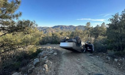 Tesla Cybertruck overturned on a narrow mountain dirt road surrounded by pine trees and rocky terrain