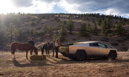 Two people near horses and hay bales beside a Tesla Cybertruck in a mountainous, forested landscape under a partly cloudy sky.