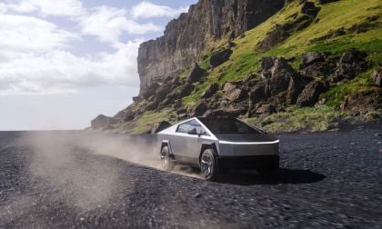 Stainless steel Tesla Cybertruck driving on rocky volcanic terrain, kicking up dust with steep cliff backdrop