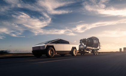 A silver Tesla Cybertruck shown from a side 3/4 angle, towing what appears to be a rocket engine on a trailer during sunset with dramatic clouds in the sky.