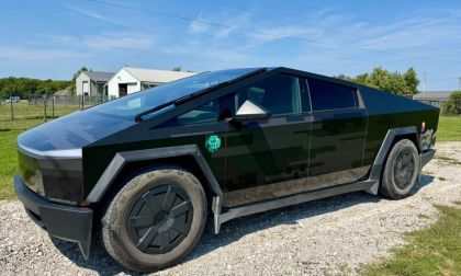 Black futuristic truck parked on gravel near a fenced grassy area and buildings.