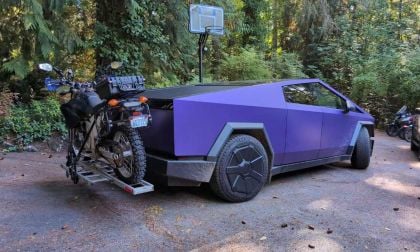 Purple futuristic truck with a motorcycle mounted on the back, parked in a wooded area.