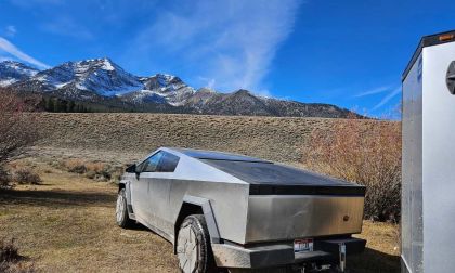 A Tesla Cybertruck in silver/stainless steel finish is parked in a mountainous landscape, shown from a rear three-quarter angle with snow-capped peaks visible in the background.