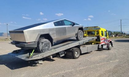 Stainless steel 2025 Tesla Cybertruck loaded on bright yellow tow truck against rural mountain backdrop