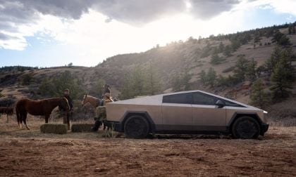 Tesla Cybertruck stainless steel electric pickup truck, side profile view, angular design, parked in rural desert landscape with horses and riders.
