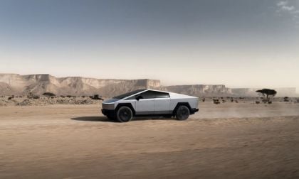 A Tesla Cybertruck drives through a dusty desert landscape, with rugged cliffs and sparse vegetation in the background.