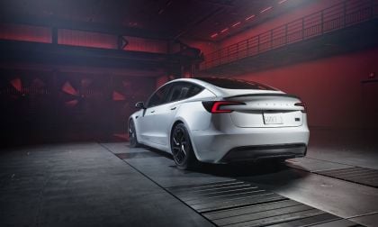 Rear three-quarter view of a white Tesla Model 3 Highland in an industrial wind tunnel testing environment with dramatic red lighting. The sleek aerodynamic design is highlighted by performance tires and minimalist rear end.