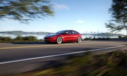 Red car driving on a road beside a lake, with a city skyline in the background.
