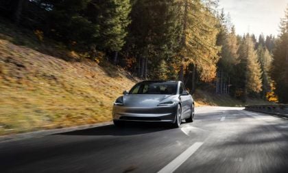 Silver Tesla Model 3 driving on winding forest road during autumn sunset