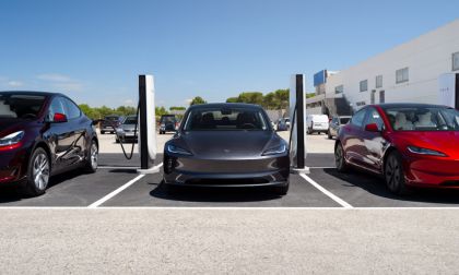 Multiple Tesla Model 3 vehicles in gray and red parked at charging stations, with white Supercharger pedestals visible, outside a modern commercial building.