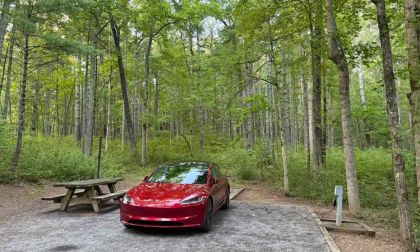 Red electric car charging at a forest campsite, surrounded by tall green trees. A wooden picnic table is on the left, evoking a serene, eco-friendly vibe.