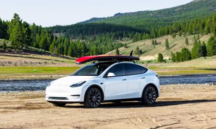 A white Tesla Model Y parked on sandy terrain by a river, with a red surfboard secured on its roof against a backdrop of green hills.