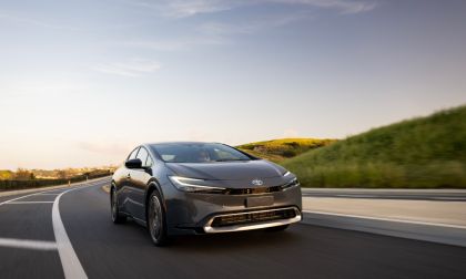 2025 Toyota Prius in red driving on a winding forest road, front three-quarter view emphasizing sporty exterior design and advanced hybrid technology.