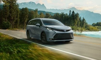Silver 2025 Toyota Sienna minivan shown from a front three-quarter view, driving on a scenic mountain road with snow-capped peaks and turquoise water in the background.