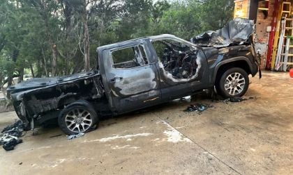 Severely fire-damaged black Toyota Tundra with melted rear section, parked outside brick building. Chrome wheels remain intact despite extensive burn damage.