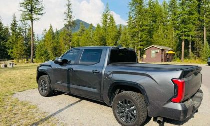 A dark gray/charcoal 2025 Toyota Tundra pickup truck shown from the rear quarter view, parked in a scenic mountain setting with evergreen trees and cabins in the background.