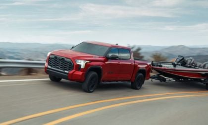 Red Toyota Tundra towing a boat on a winding mountain road. The backdrop shows a scenic landscape of rolling hills under a partly cloudy sky.