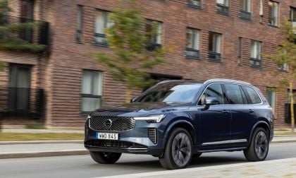 A 2025 blue Volvo CX90, speeding past modern brick buildings, showcasing its stylish design and dynamic performance.
