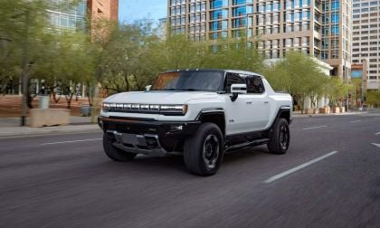 A white 2025 GMC Hummer EV drives through a city street with modern buildings and greenery in the background, showcasing its rugged design.