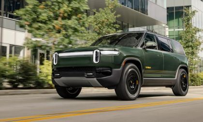 Green Rivian R1S electric SUV driving through a modern city street with glass office buildings in the background
